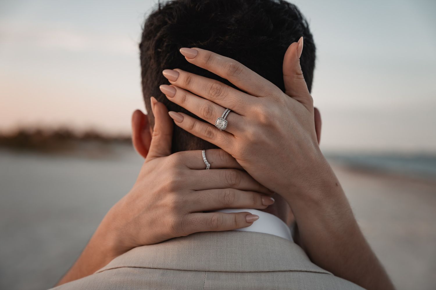 Close-up of bride's hand with wedding ring resting on groom's neck at beach.