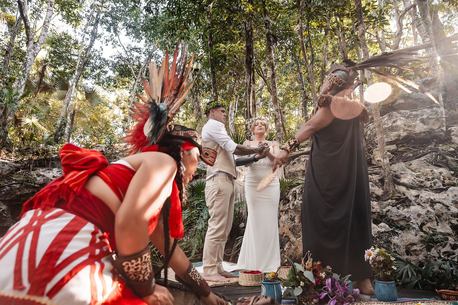 Outdoor jungle wedding ceremony with couple holding hands, officiant, and performer in traditional red feathered costume.