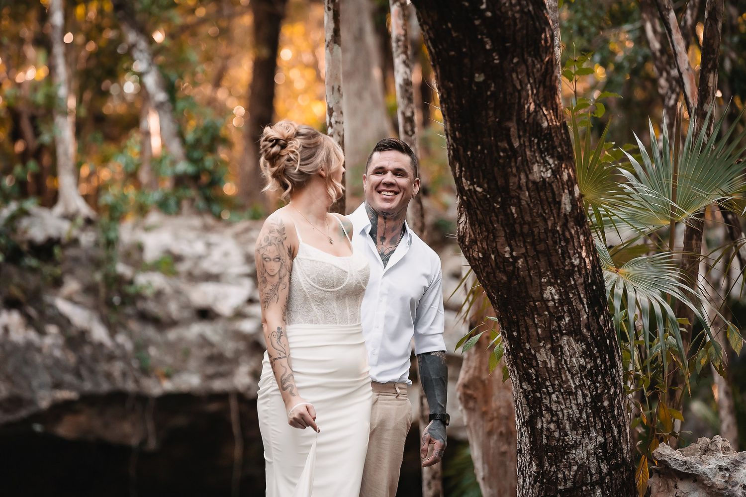 Couple in wedding attire smiling together in a lush forest setting, bride in white gown with tattoos, groom in white shirt.