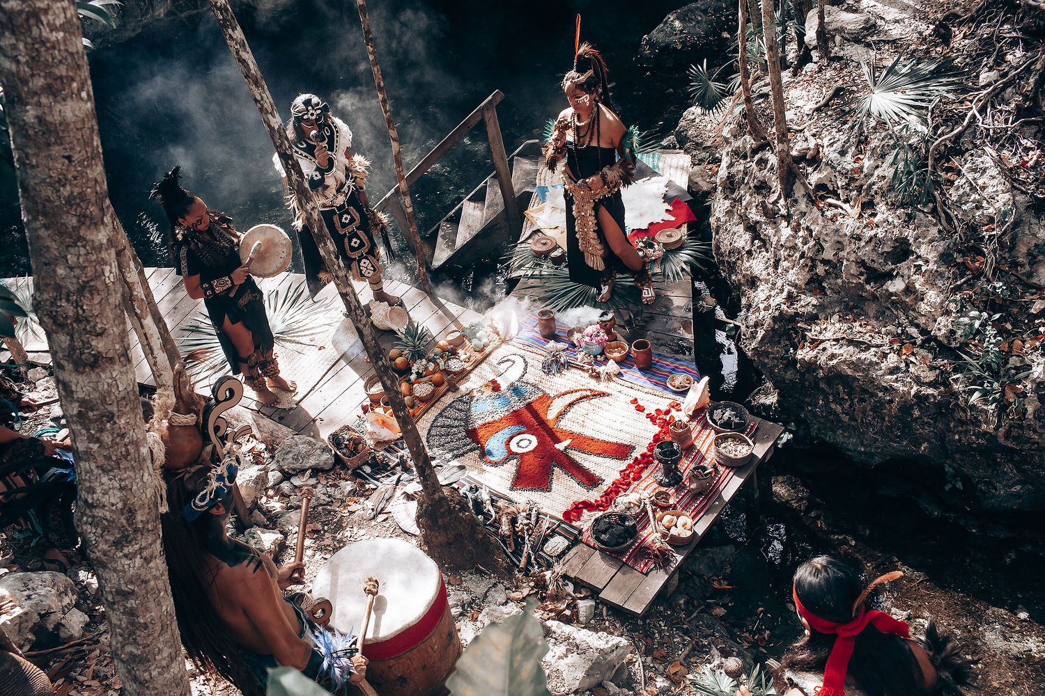 Musicians in traditional dress perform with drums and instruments during a cultural celebration in a forest setting.