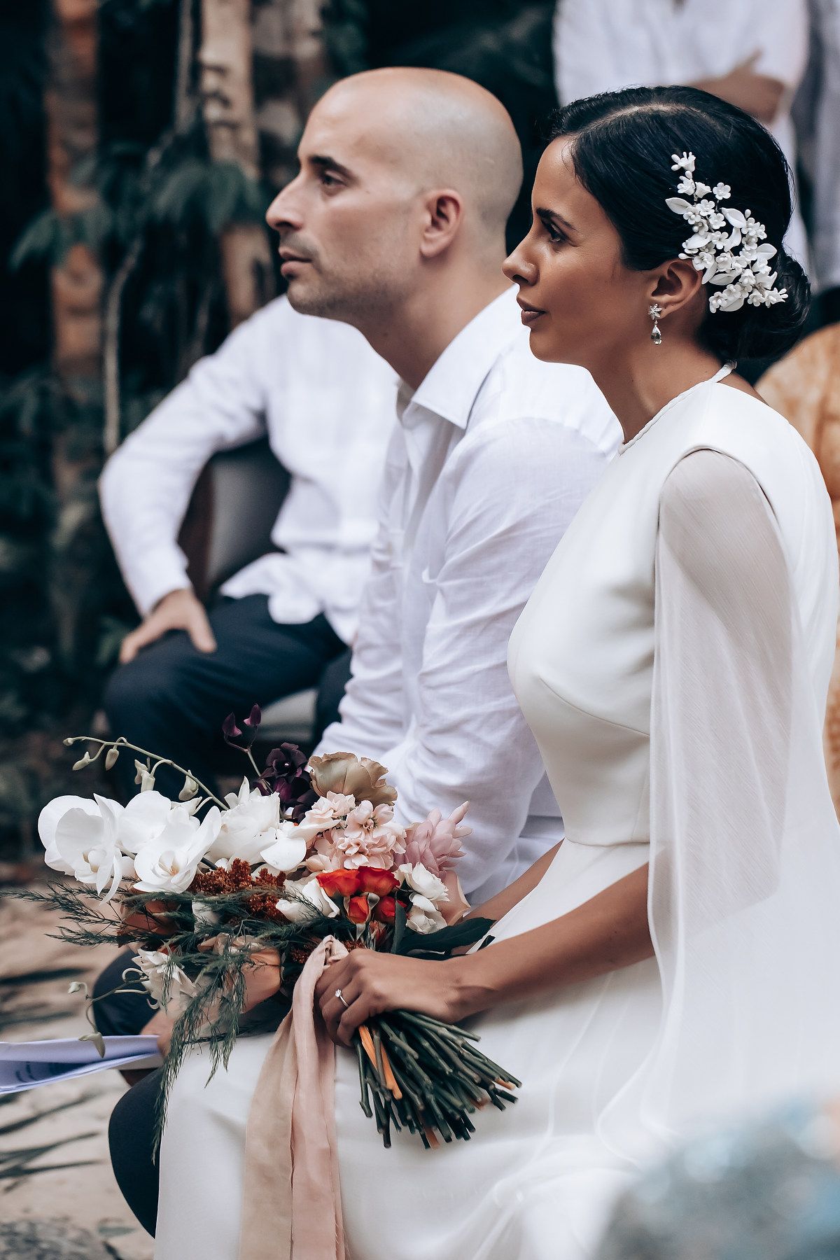 Wedding ceremony guests in white attire seated outdoors holding floral bouquets during daytime celebration.