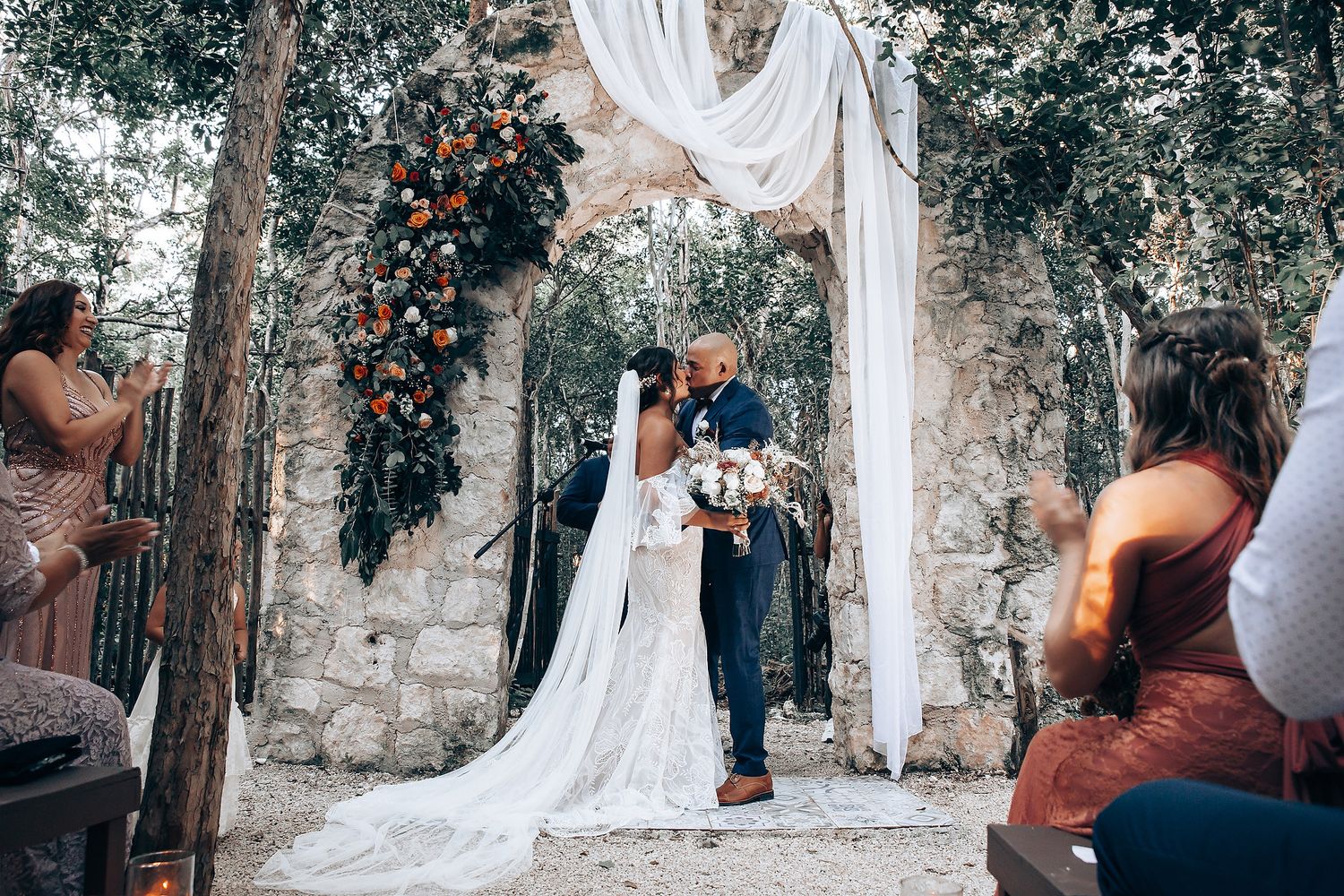 Newlyweds stand beneath an elegant white draped arch decorated with orange flowers.