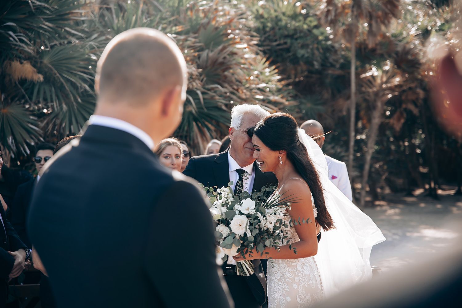 A romantic beach wedding ceremony with guests seated in white chairs against an ocean backdrop.