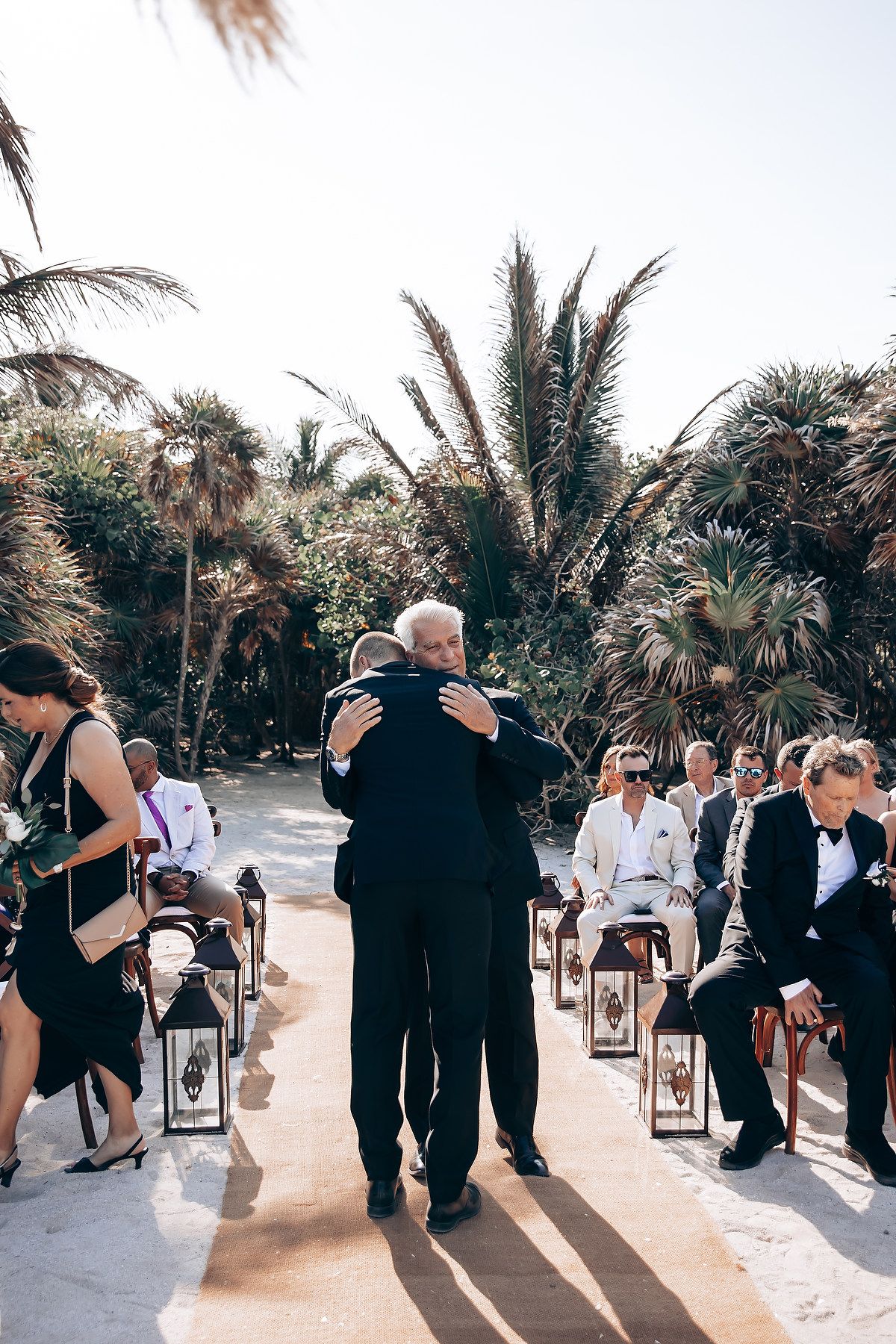 Wedding guests gathering on a sunlit beach path lined with palm trees.