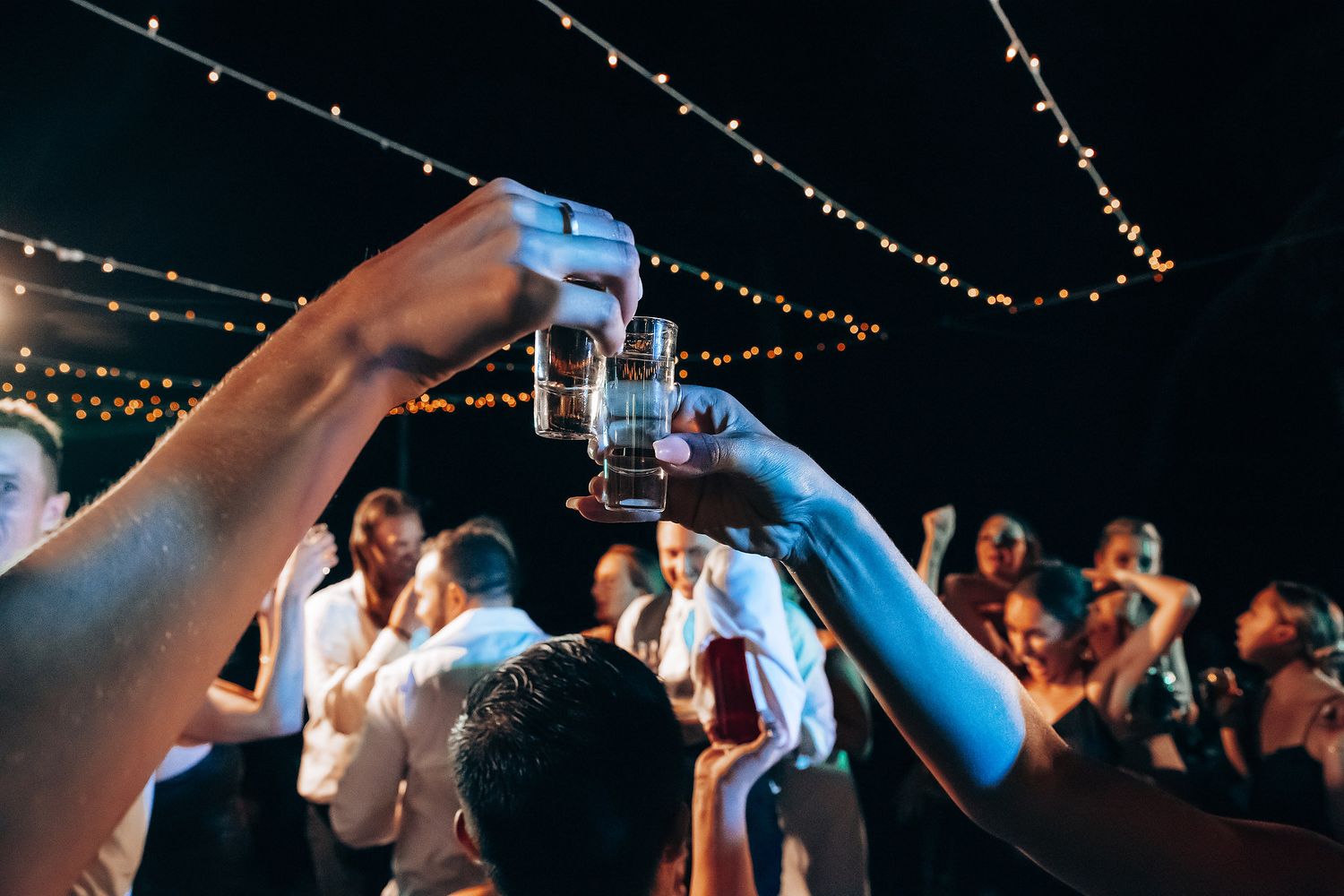 String lights illuminate a dark reception venue as guests dance and celebrate together.