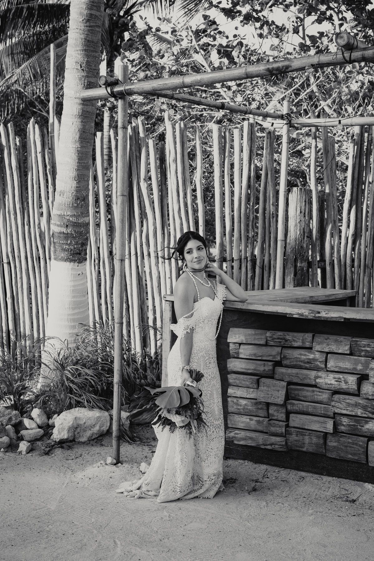 Elegant bride in lace gown holding a bouquet poses by rustic bamboo fence on sandy beach in black and white photo.