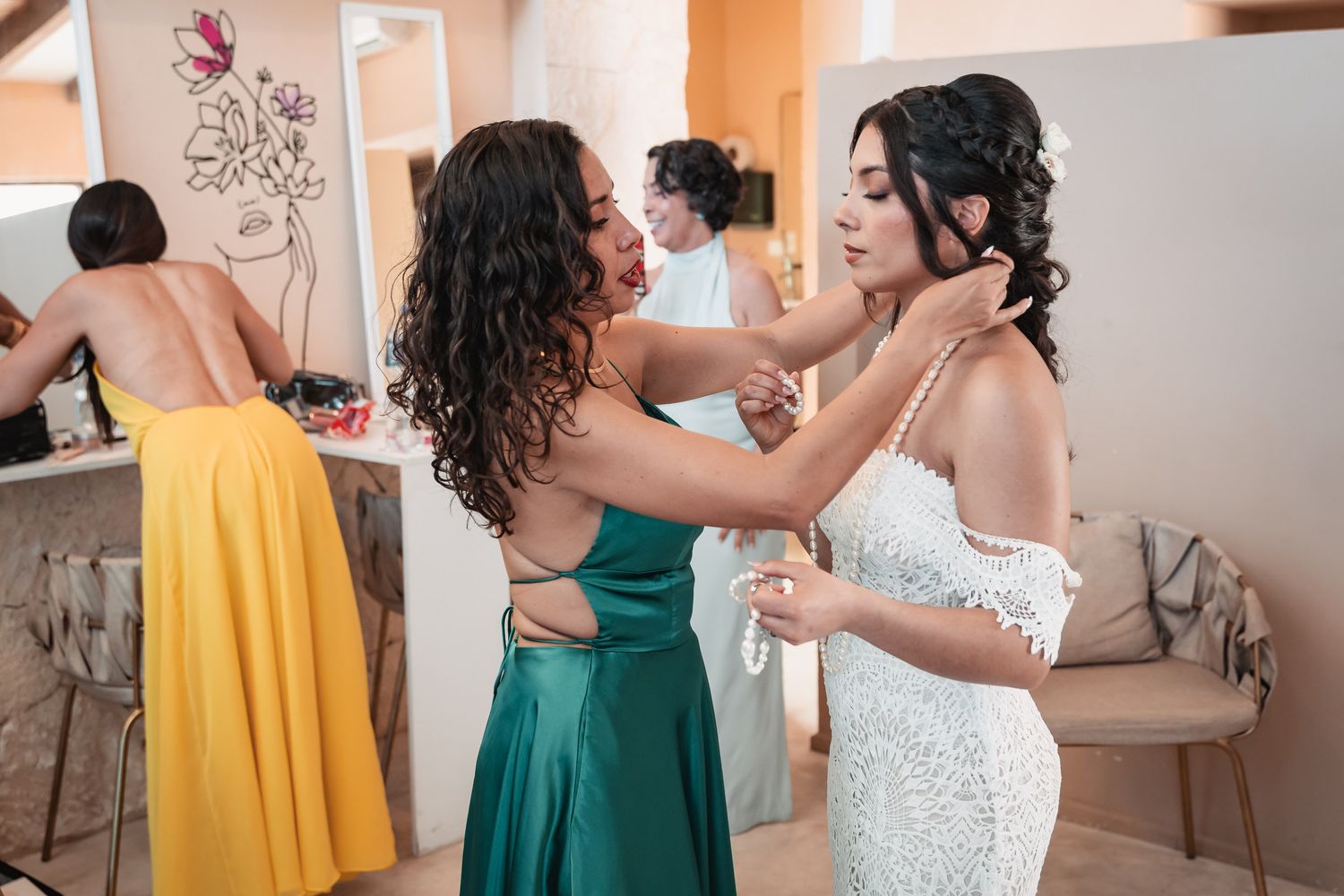 A bridesmaid in green helps the bride with her necklace while another bridesmaid in yellow stands nearby.