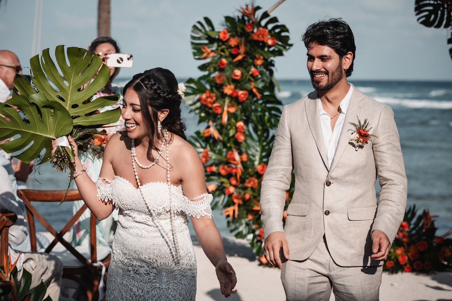 Happy bride and groom walking together after beach wedding ceremony with tropical orange floral arch backdrop.