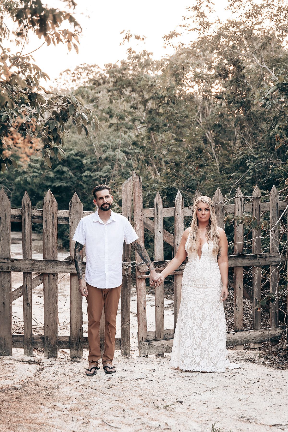 A couple stands together in front of a rustic wooden fence in an outdoor garden setting.