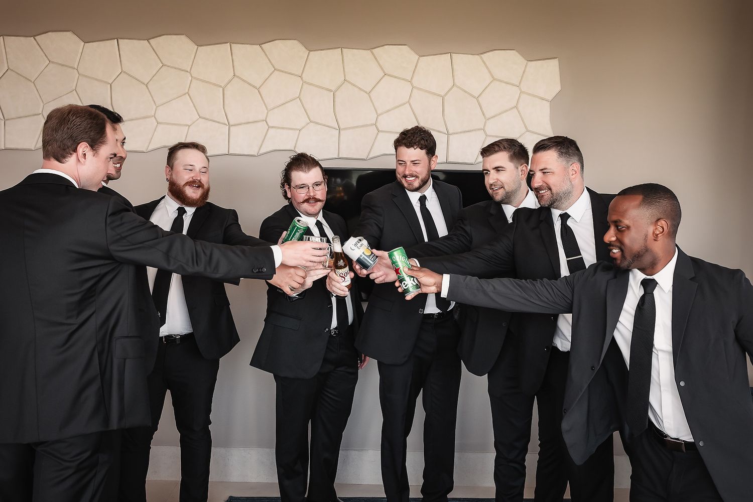 Wedding party in black suits posing together against a beige wall for a fun group photo.