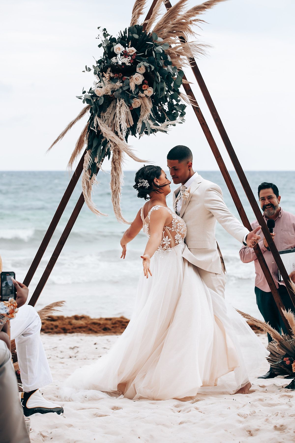 Wedding couple sharing a moment under floral arch with pampas grass on beach.