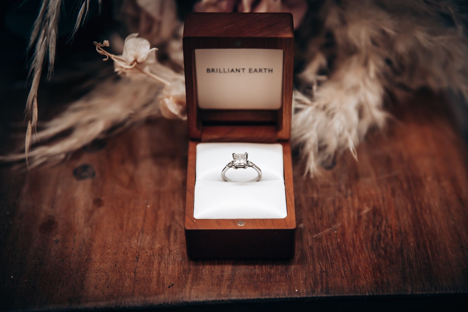 A wooden ring box with delicate white feathers sits on a dark rustic wooden surface.