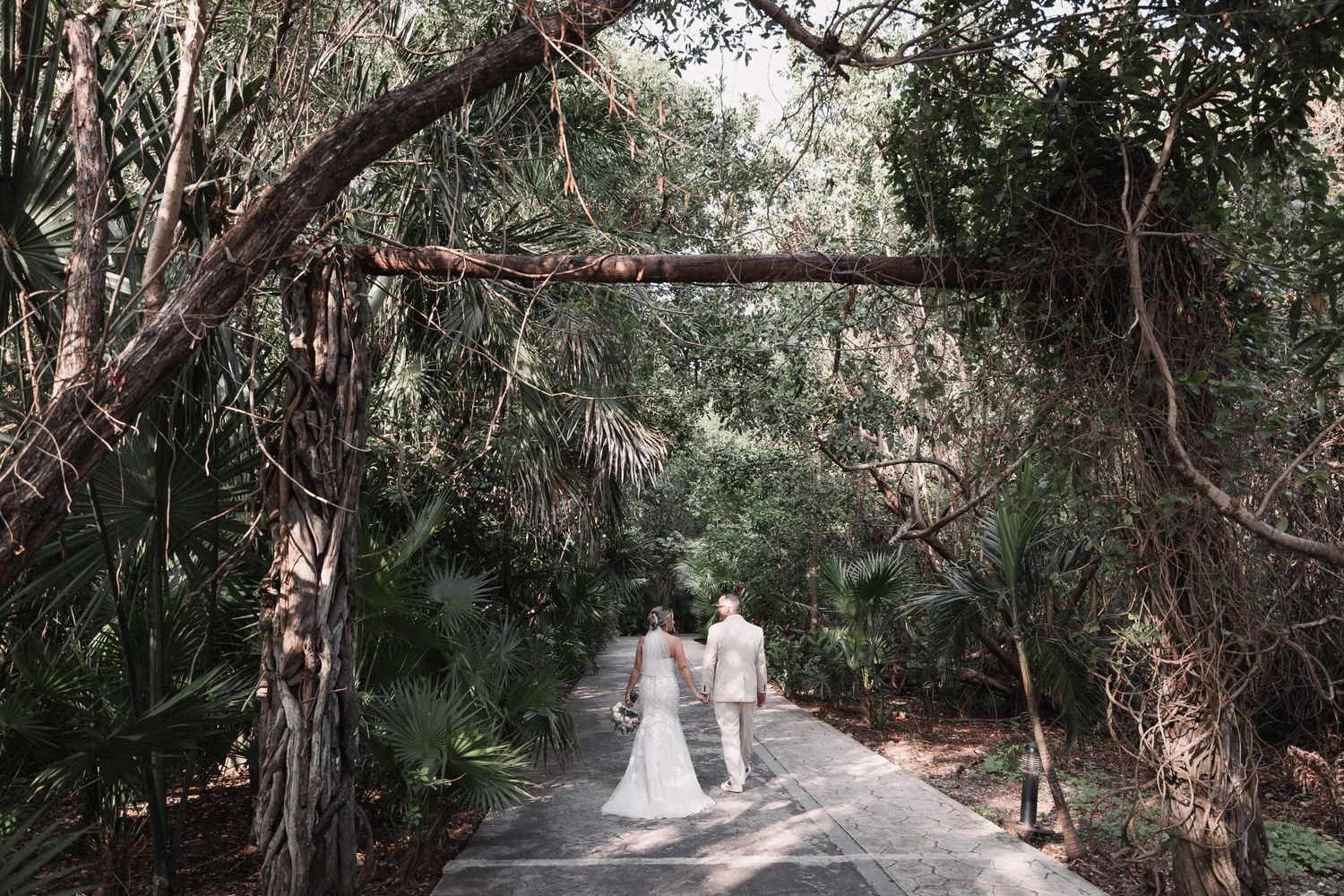 Bride and groom walk hand in hand down lush tropical pathway beneath natural tree archway.