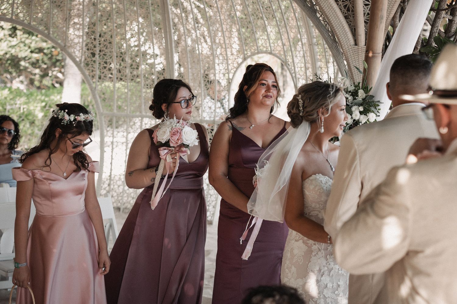 Bridesmaids in mauve and dusty rose gowns standing at outdoor beach wedding ceremony.