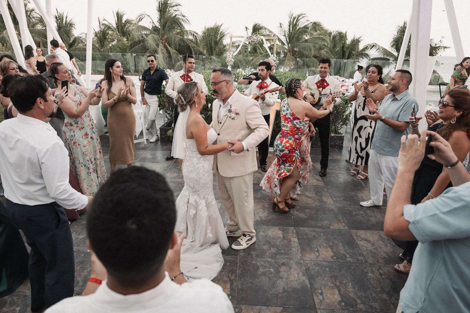 Bride and groom share first dance surrounded by cheering guests at outdoor tropical wedding reception.