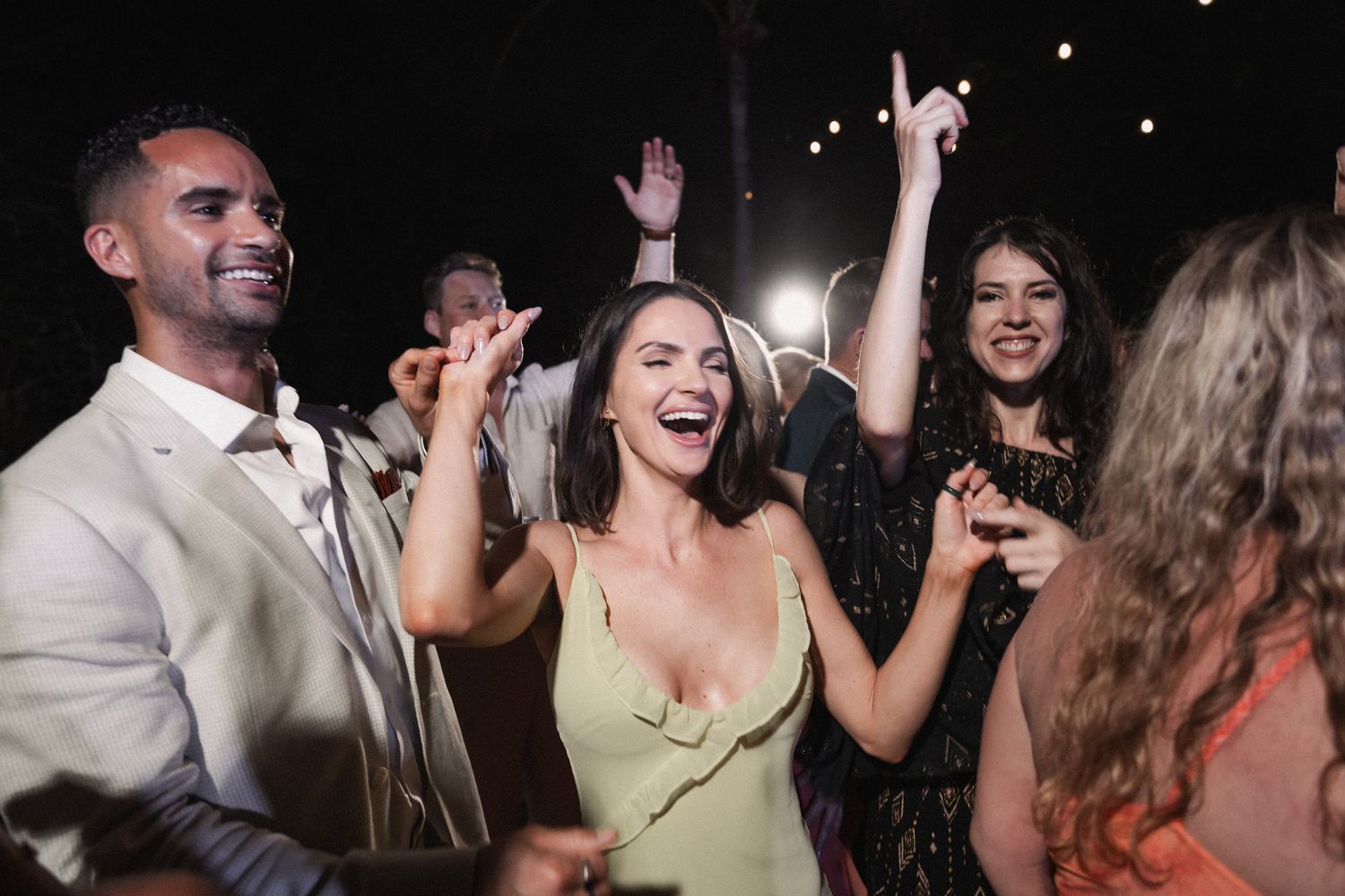 Wedding guests dance and celebrate joyfully at lively outdoor tropical wedding reception at night.