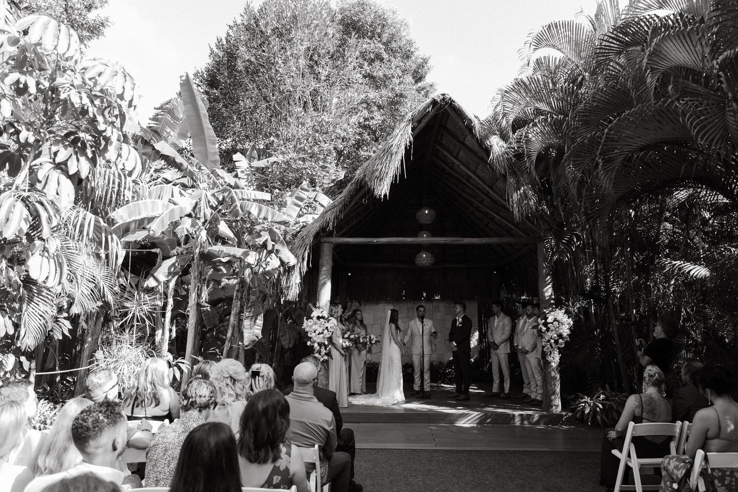 Black and white wide shot of outdoor tropical wedding ceremony with seated guests.