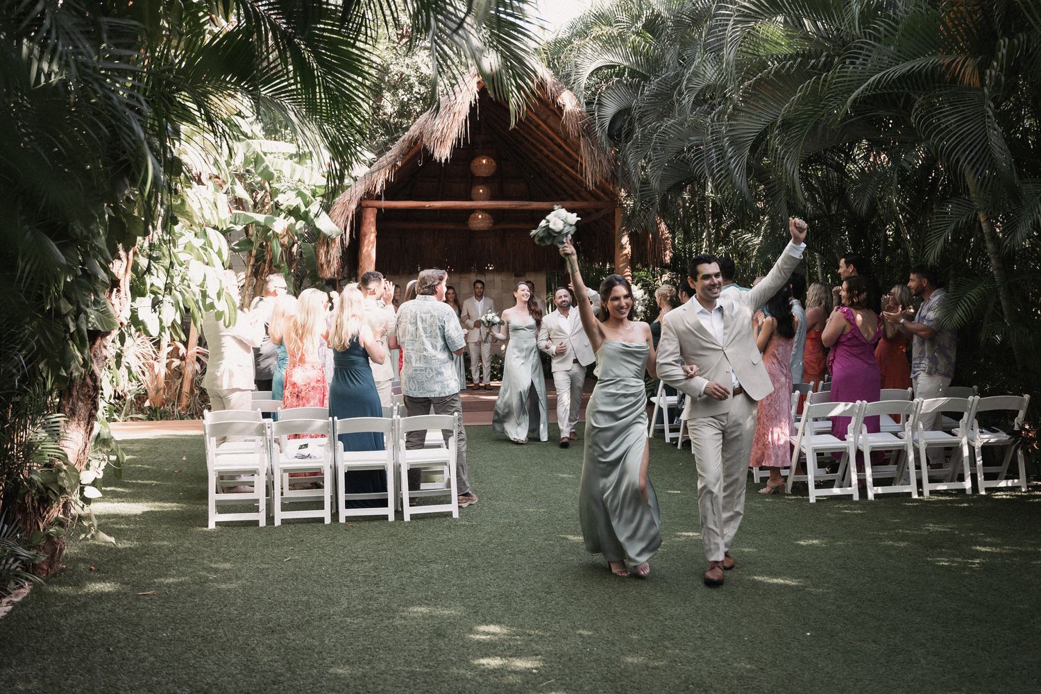 Joyful newlyweds walk down the aisle at an outdoor tropical wedding ceremony with white chairs and lush greenery.