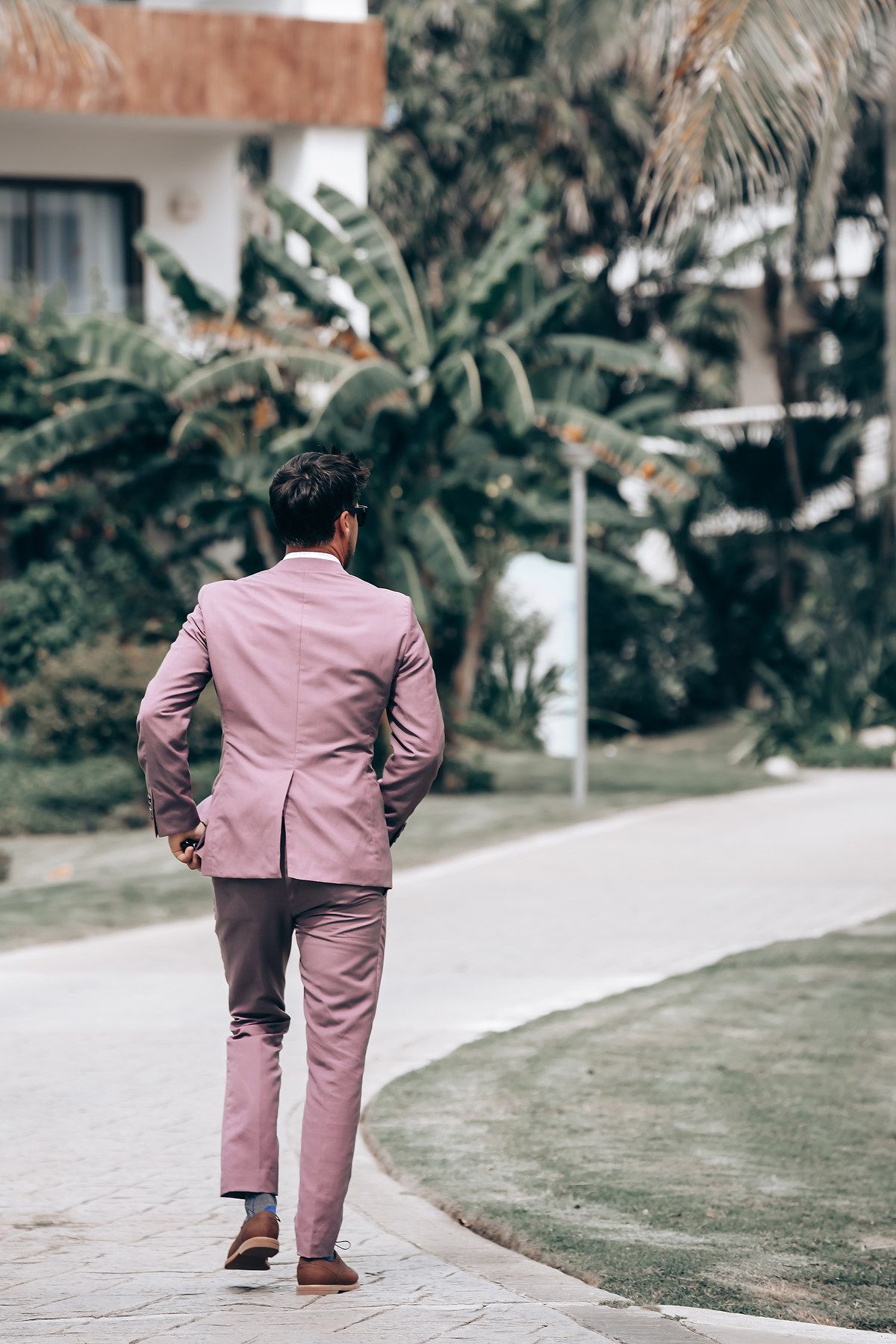Person wearing dusty pink suit walking on garden path surrounded by tropical plants.