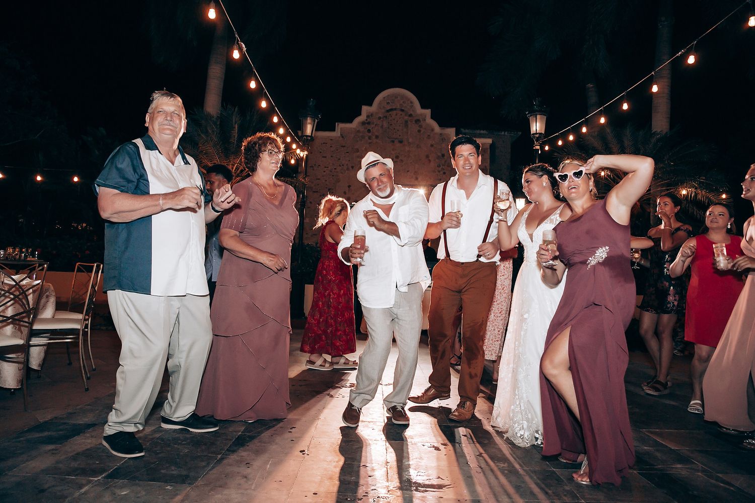 Wedding guests dancing and posing together under string lights at an outdoor evening celebration.
