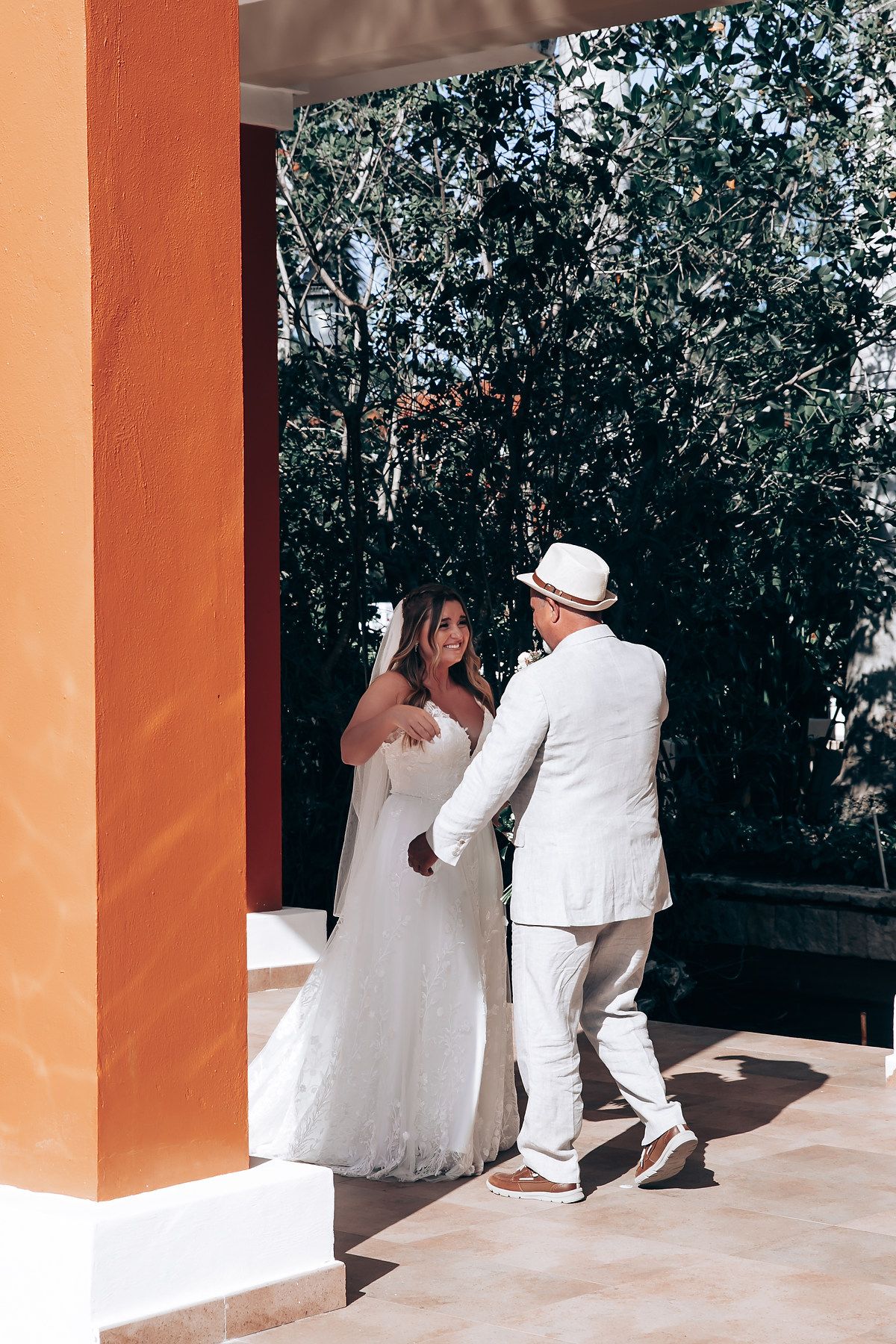 A couple in white attire poses against coral-colored pillars at an outdoor venue.