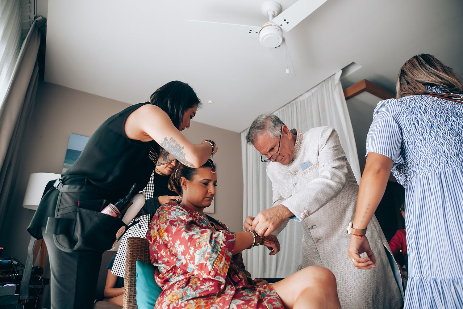 Indoor wedding preparation scene with multiple people helping with traditional dress and accessories.