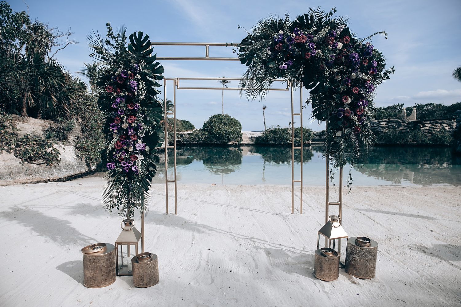 Beach wedding ceremony setup with rustic wooden arch and lanterns on white sand.
