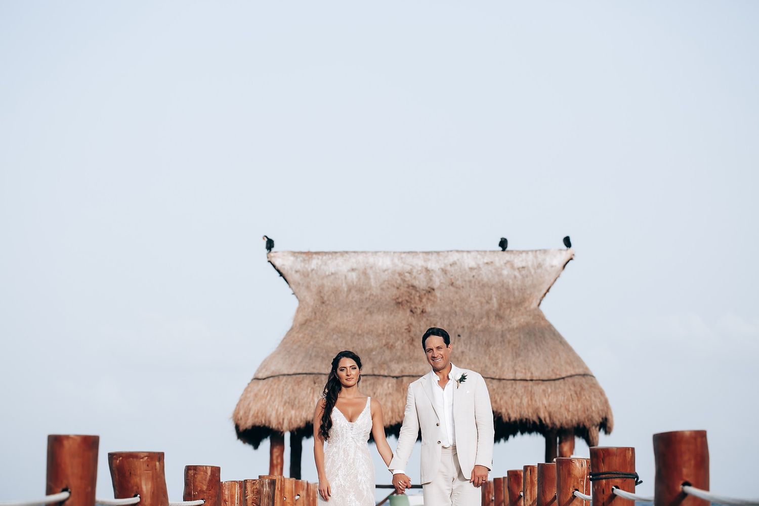 A couple poses together on a beach with palm-thatched huts and wooden posts in the background at sunset.