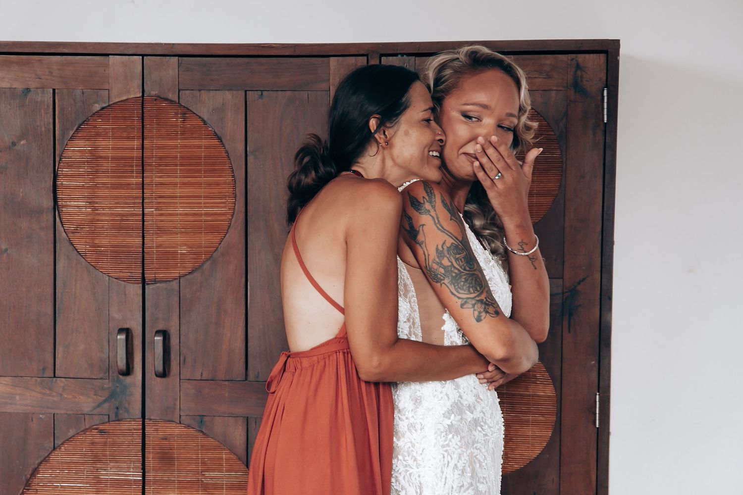Two women share an emotional moment in front of a wooden cabinet with circular designs.