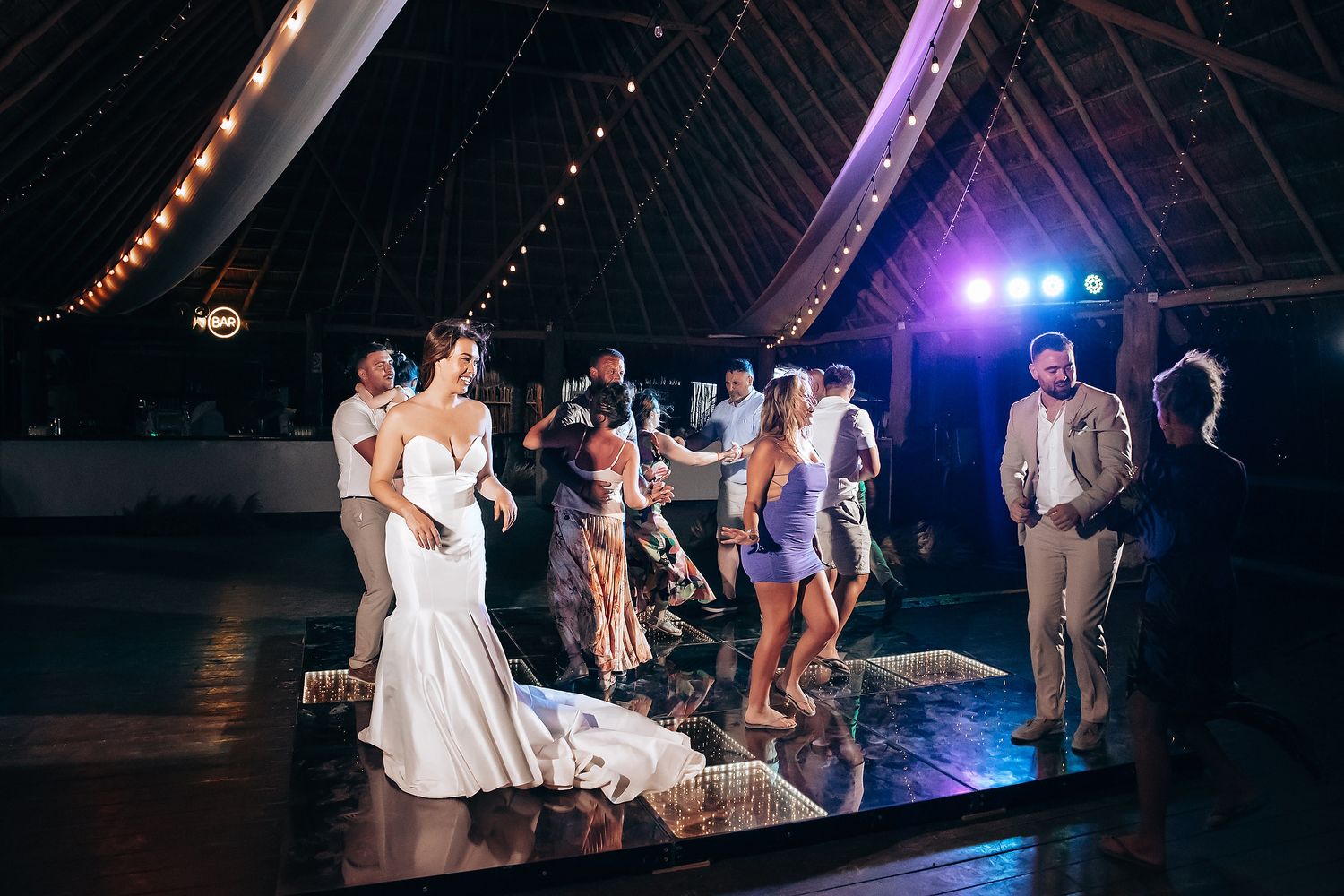 Wedding guests dance together on a dimly lit dance floor with colorful purple and blue lighting at a reception celebration.