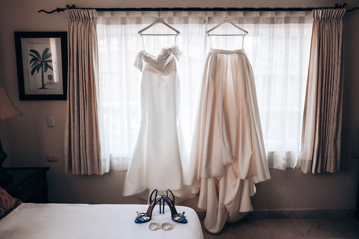 White wedding dresses hanging in a sunlit room with curtains and decorative wall art.