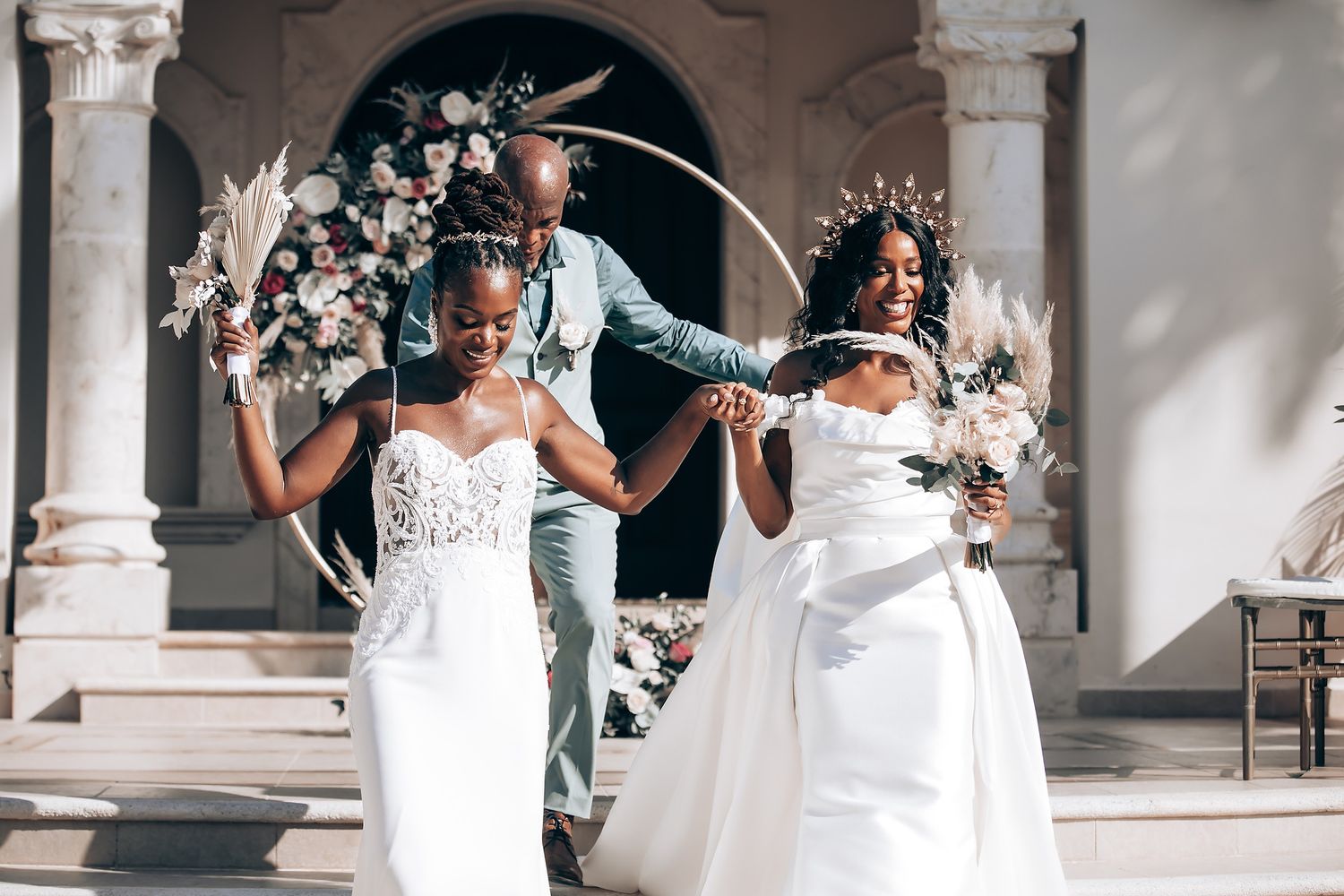 Wedding celebration with floral arch and two people in white dresses posing joyfully.
