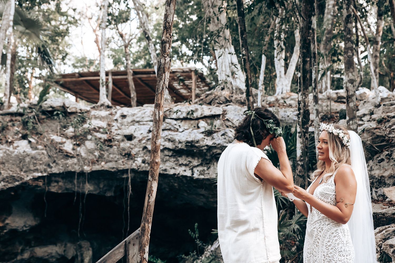A couple in white wedding attire shares romantic moments in a forest setting with natural stone formations.