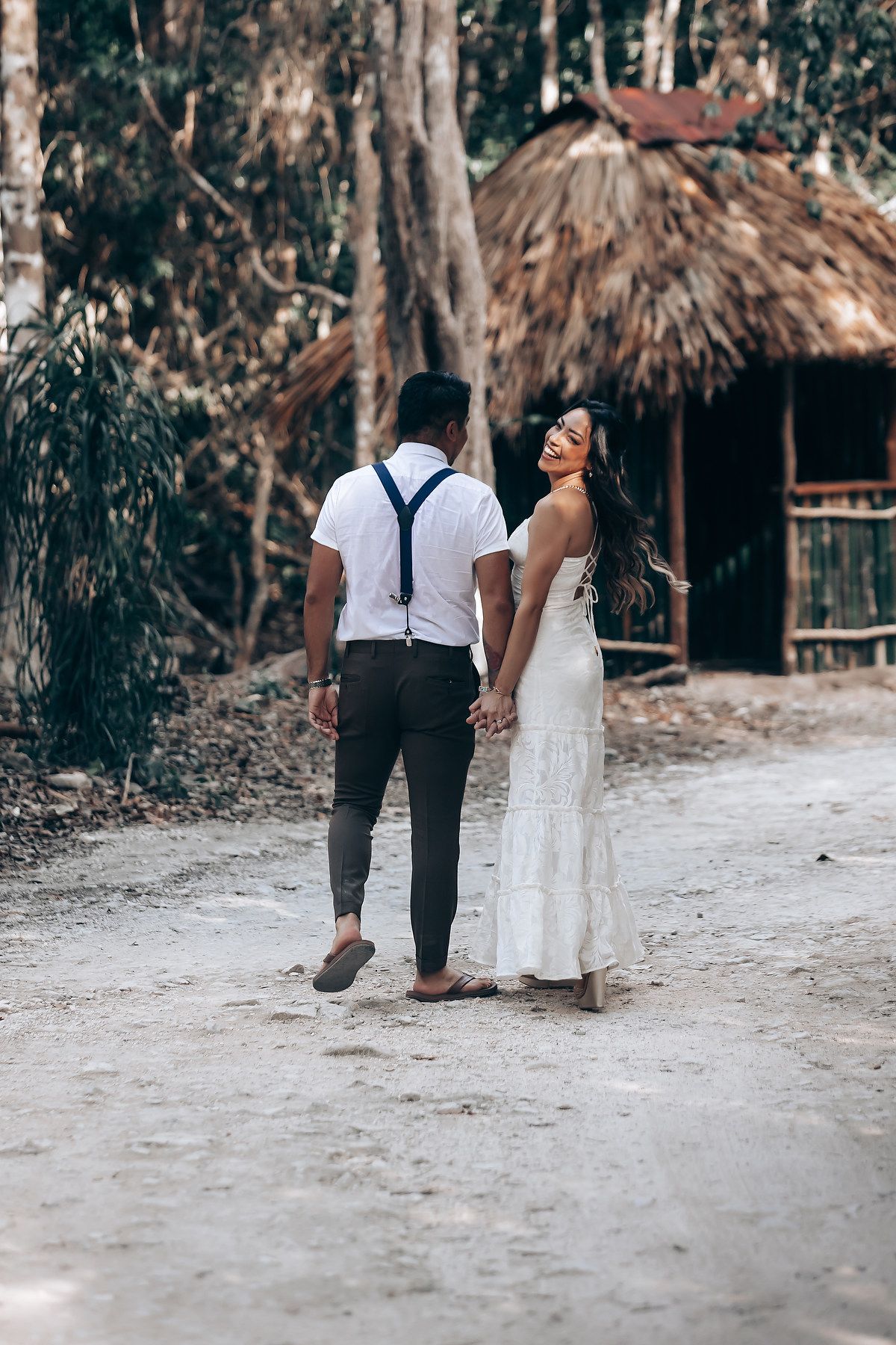 A couple walking together on a sandy path through tropical foliage wearing white and black formal attire.