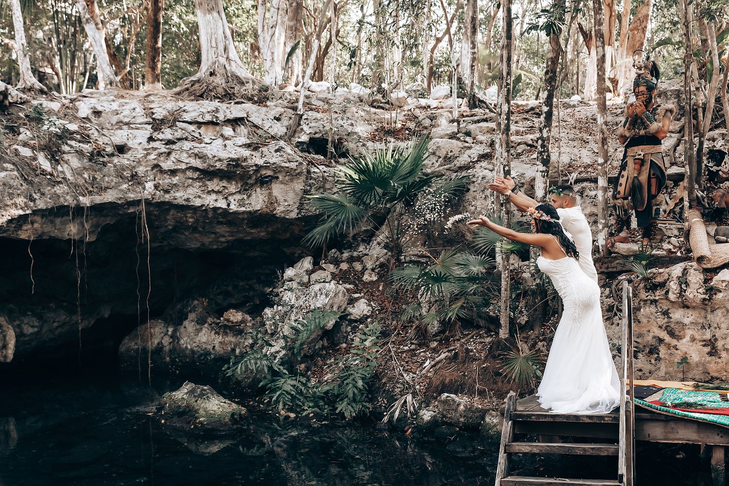 Bride in white dress stands on wooden platform overlooking natural cenote.