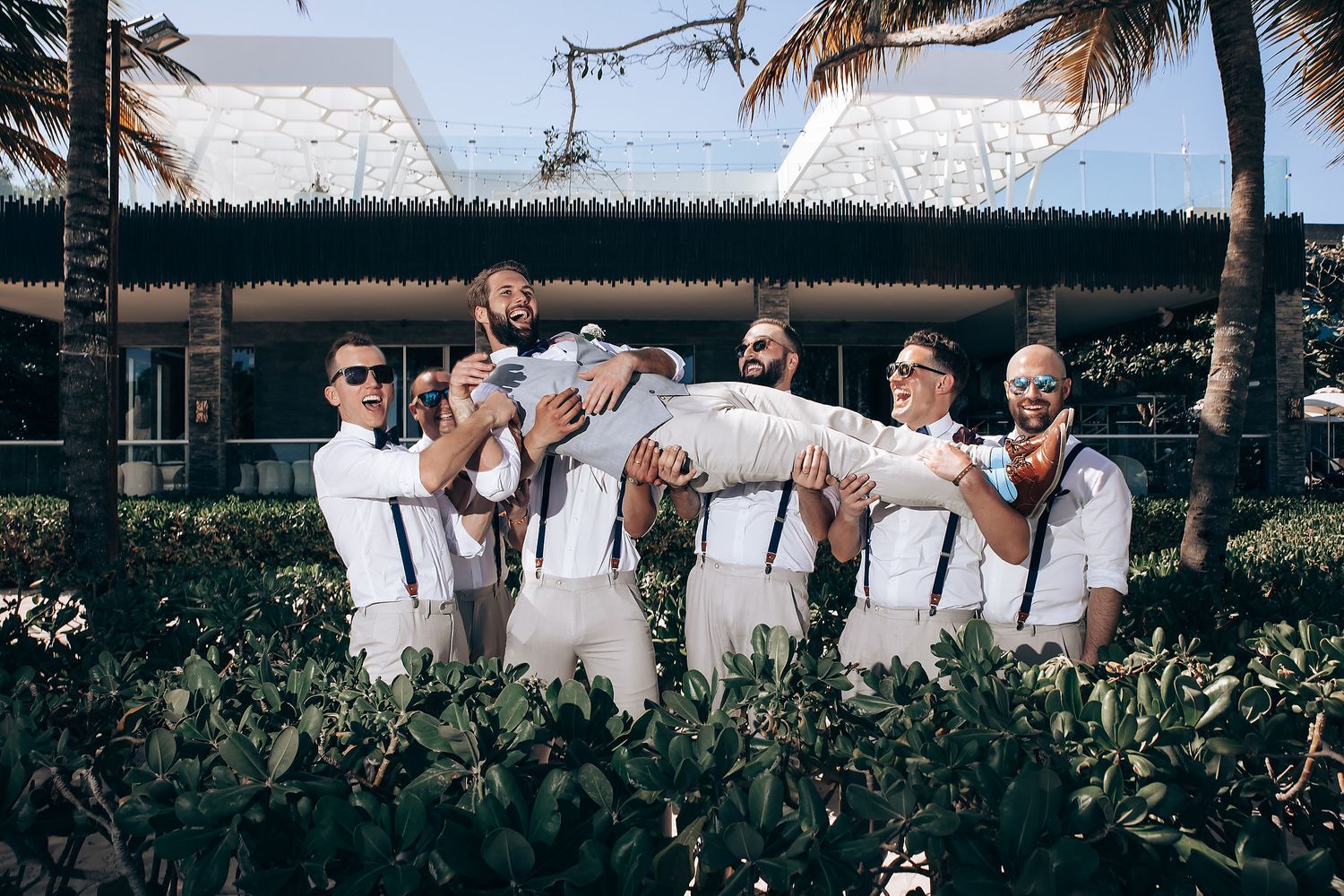 Wedding party in white attire celebrates outdoors under palm trees and blue sky.
