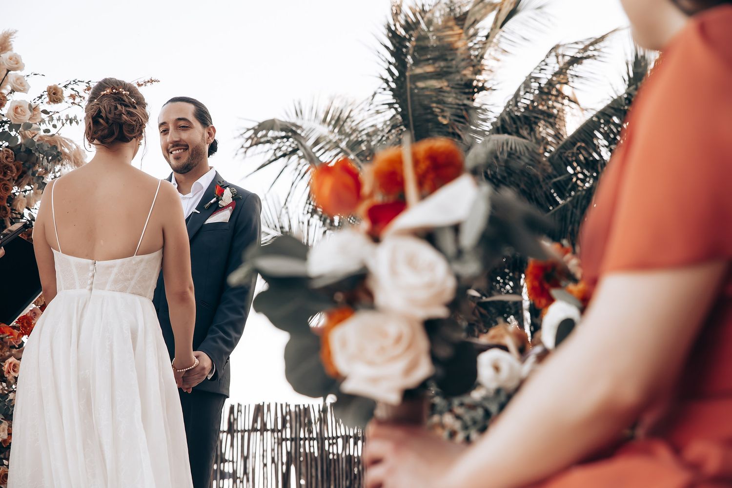 A wedding couple embraces during their outdoor ceremony as guests look on through decorative floral arrangements.