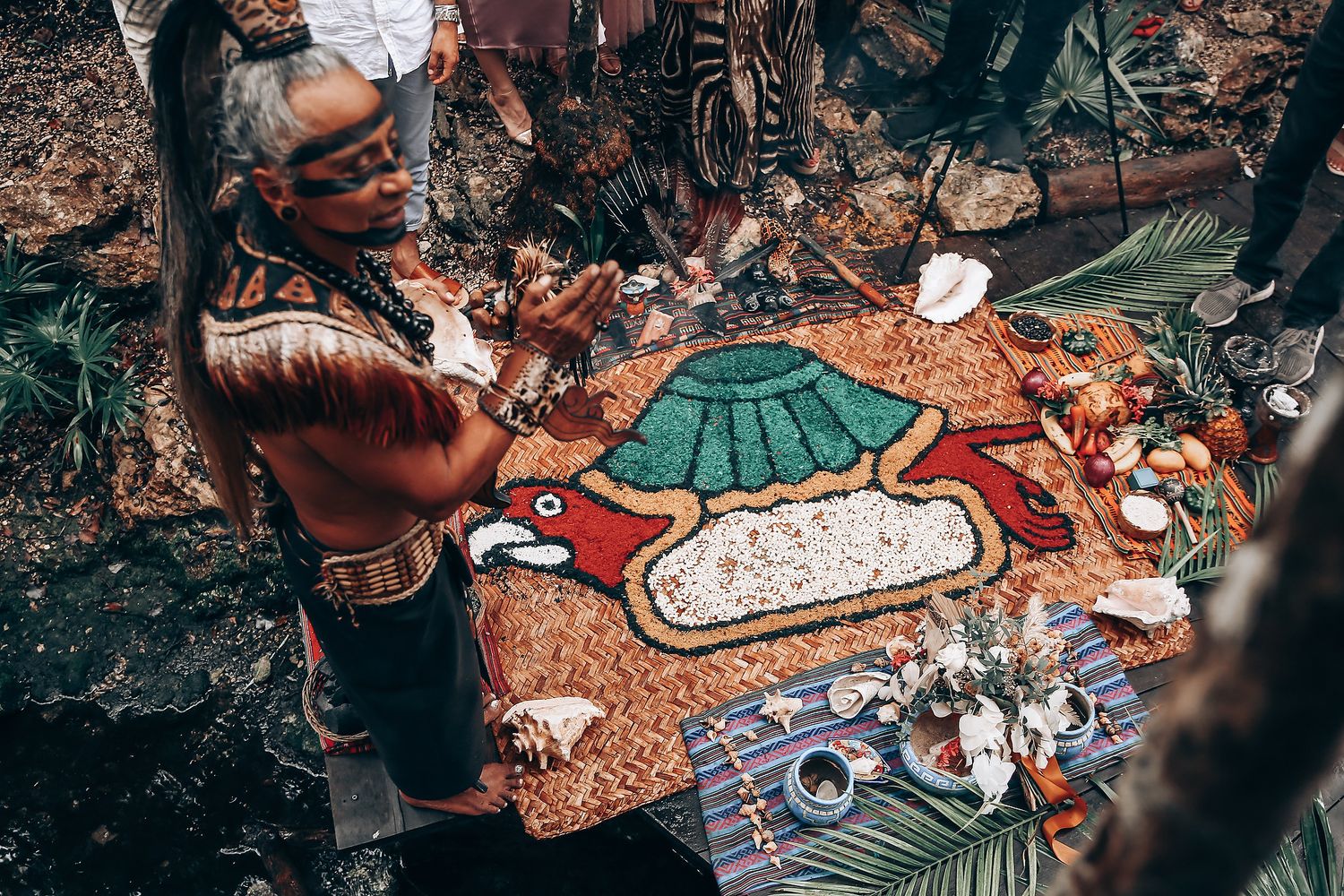 People gather around a picnic blanket with food and drinks spread out in an outdoor setting during a social gathering.