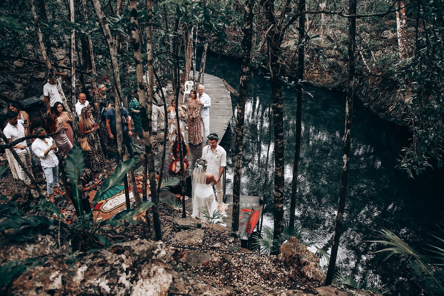 A group gathers for a traditional ceremony along a forest path with white-clad participants among the trees.