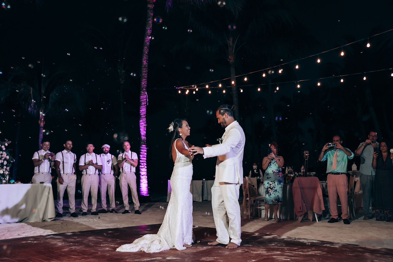 String lights illuminate an outdoor dance floor as wedding guests gather for evening festivities.