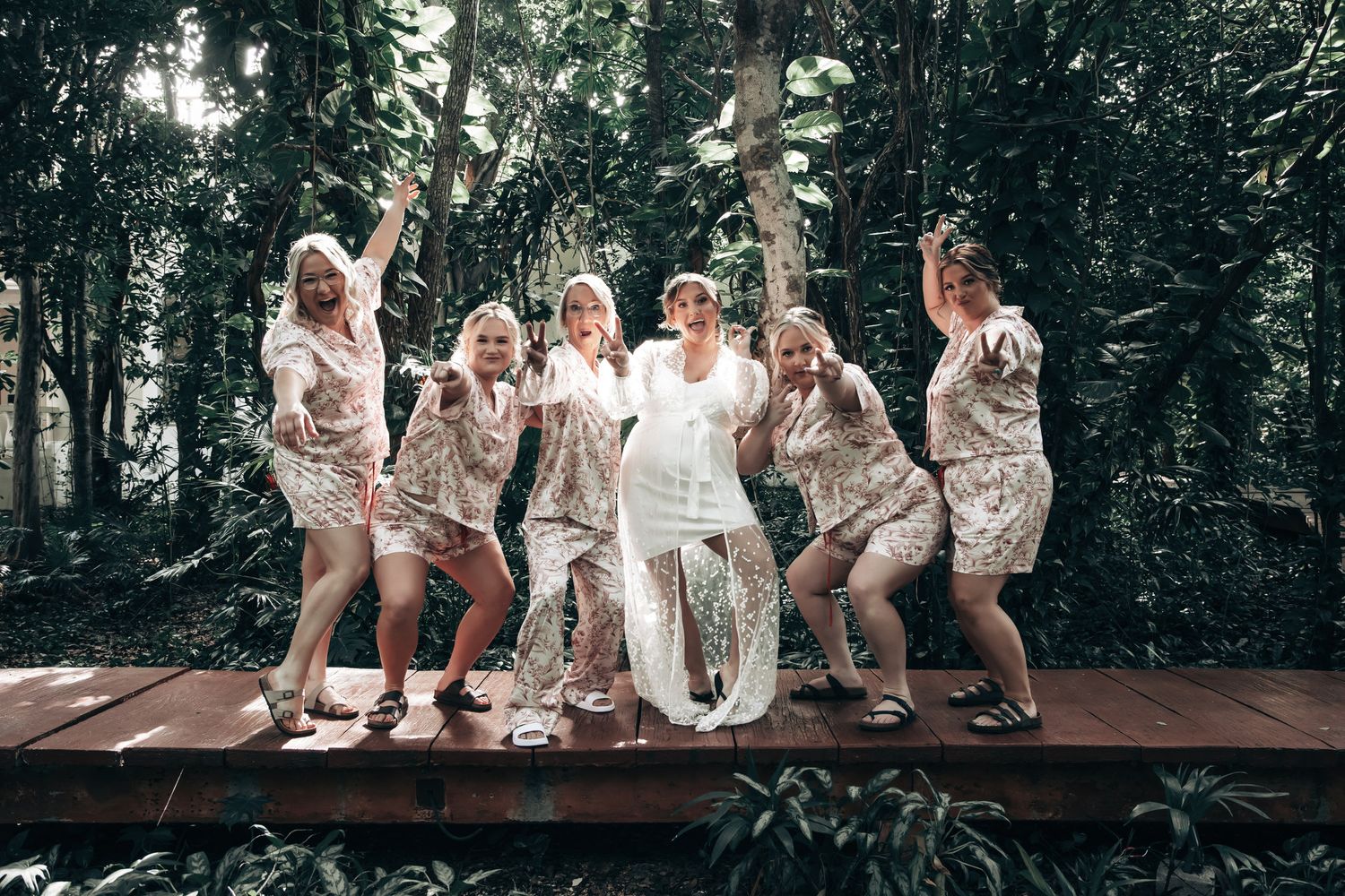 Bride and bridesmaids in matching robes celebrating on a wooden deck surrounded by lush tropical greenery.