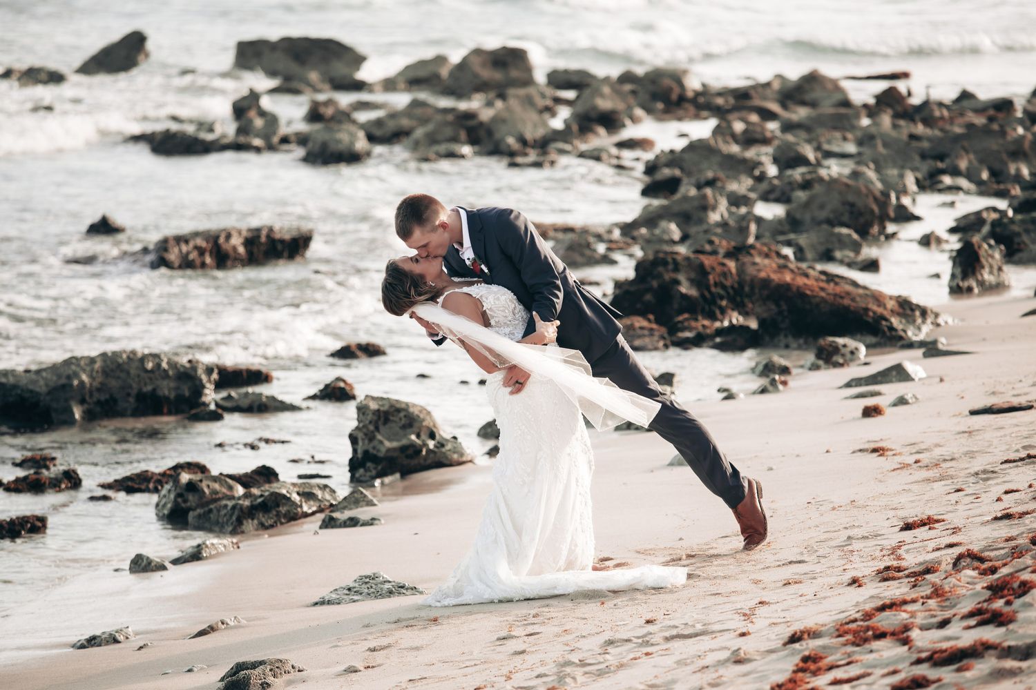 Groom dips bride for a romantic kiss on rocky ocean beach during wedding photo session.
