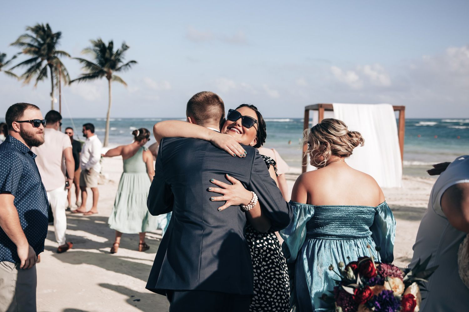 Wedding guests hugging and celebrating after beach ceremony with palm trees and ocean.