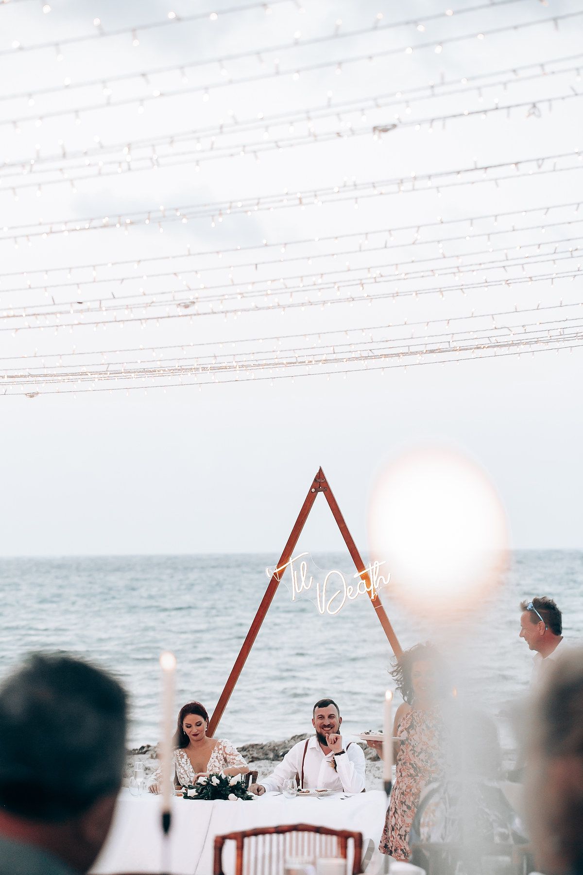 Guests celebrating at an outdoor beach wedding reception with string lights creating a romantic atmosphere.