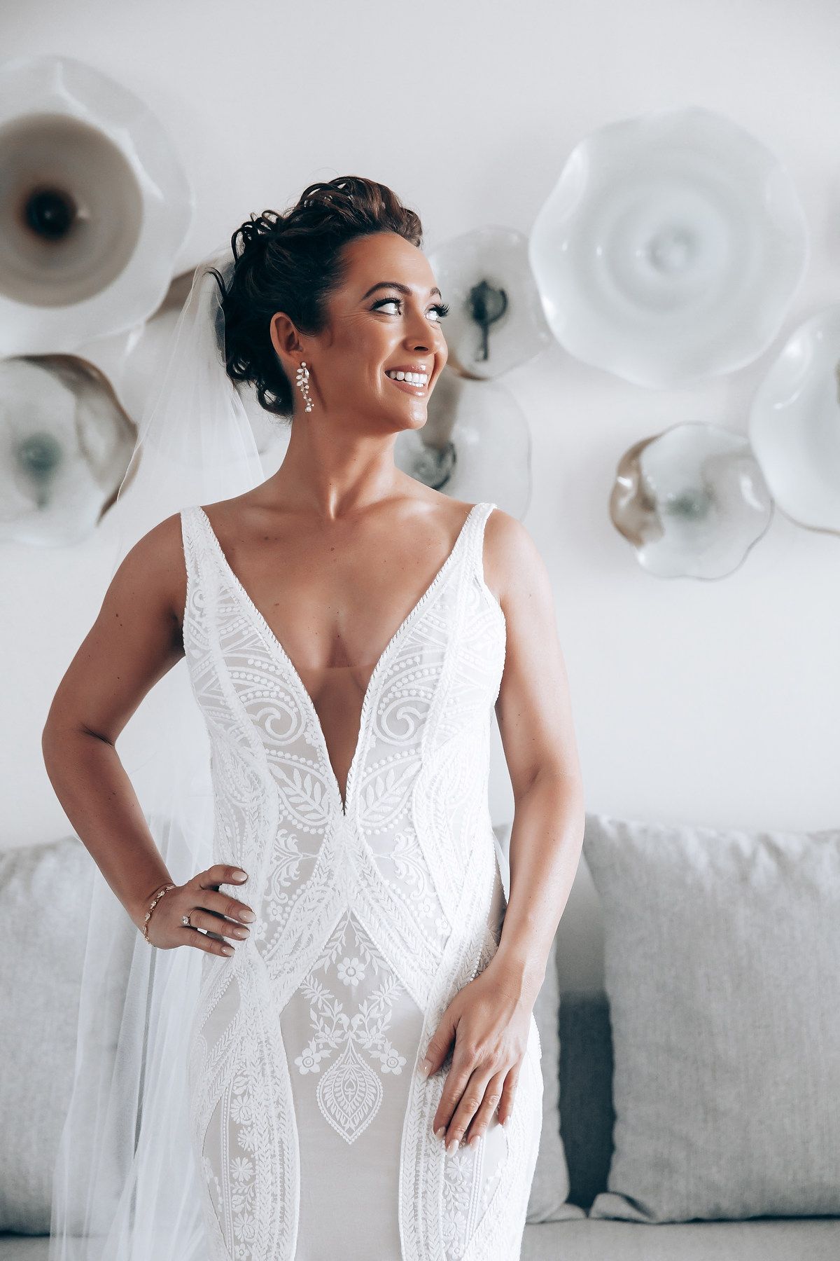 Bride in elegant white lace wedding dress poses joyfully against white decor background.