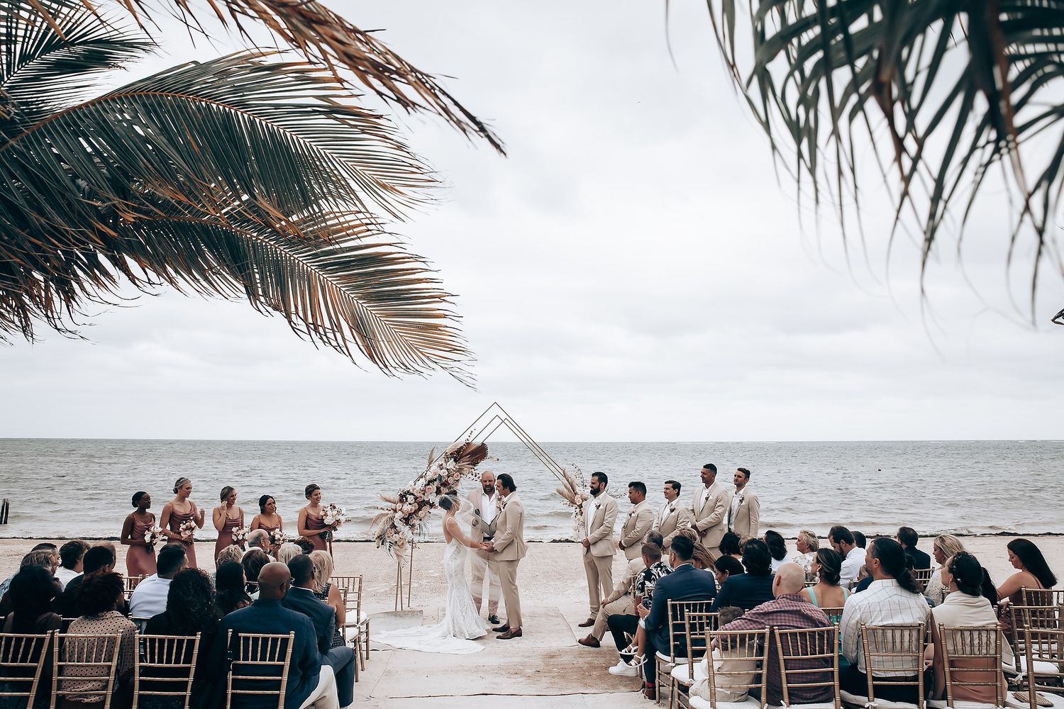 Palm fronds frame a beachside wedding ceremony with guests seated in rows.