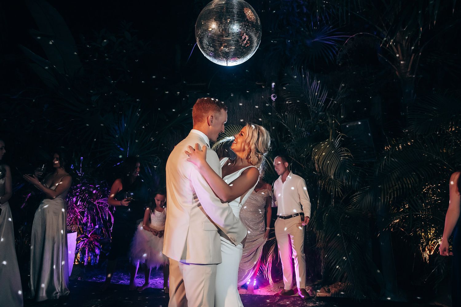 Wedding party dancing under a disco ball with purple lighting effects at night.