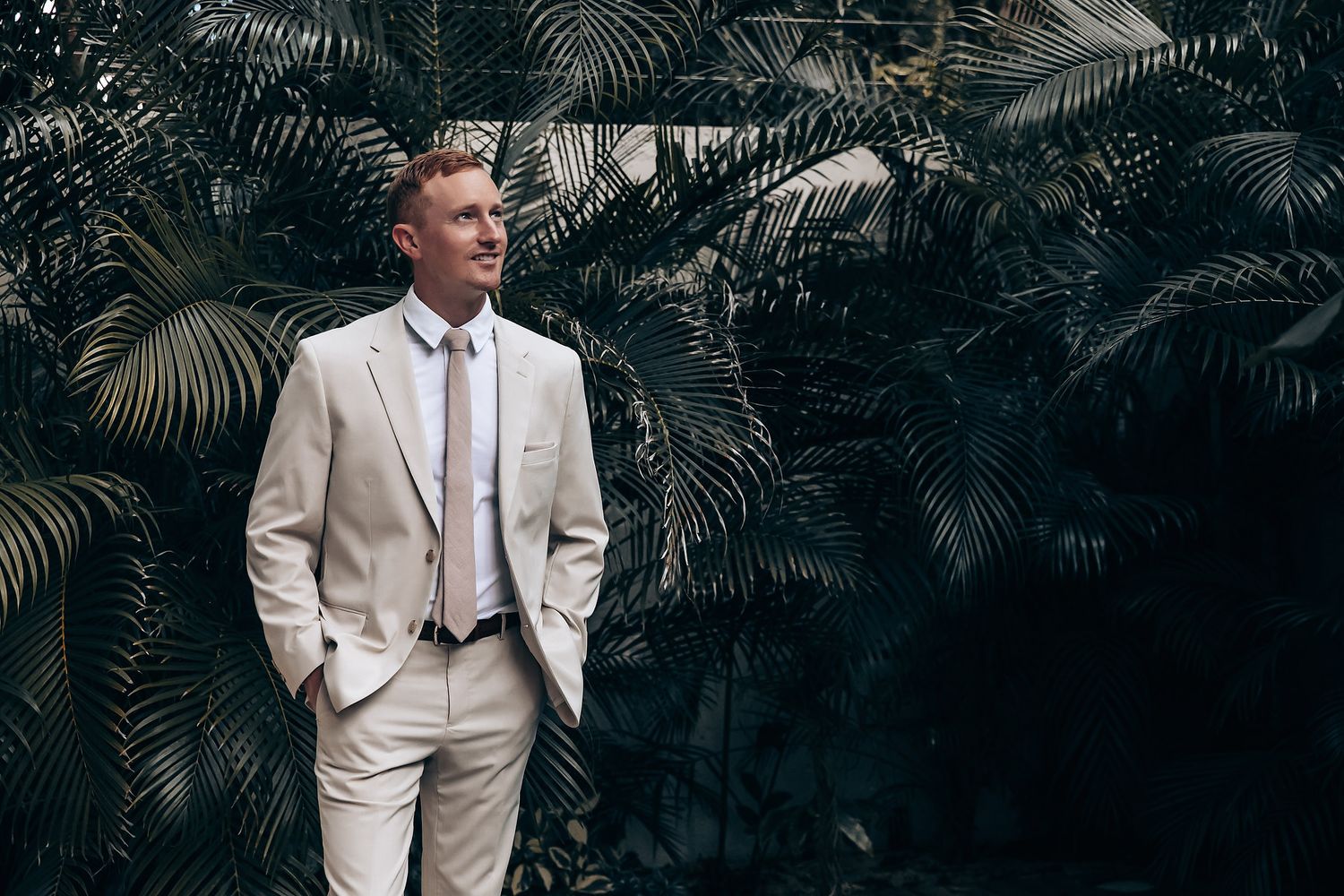 A man in a light beige suit stands confidently against a lush tropical palm background.