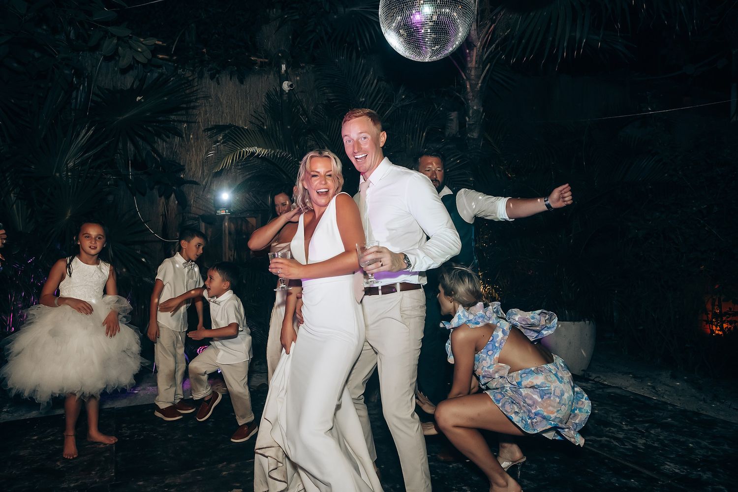 Wedding reception dance party with disco ball overhead as couple dances surrounded by guests on dark dance floor.