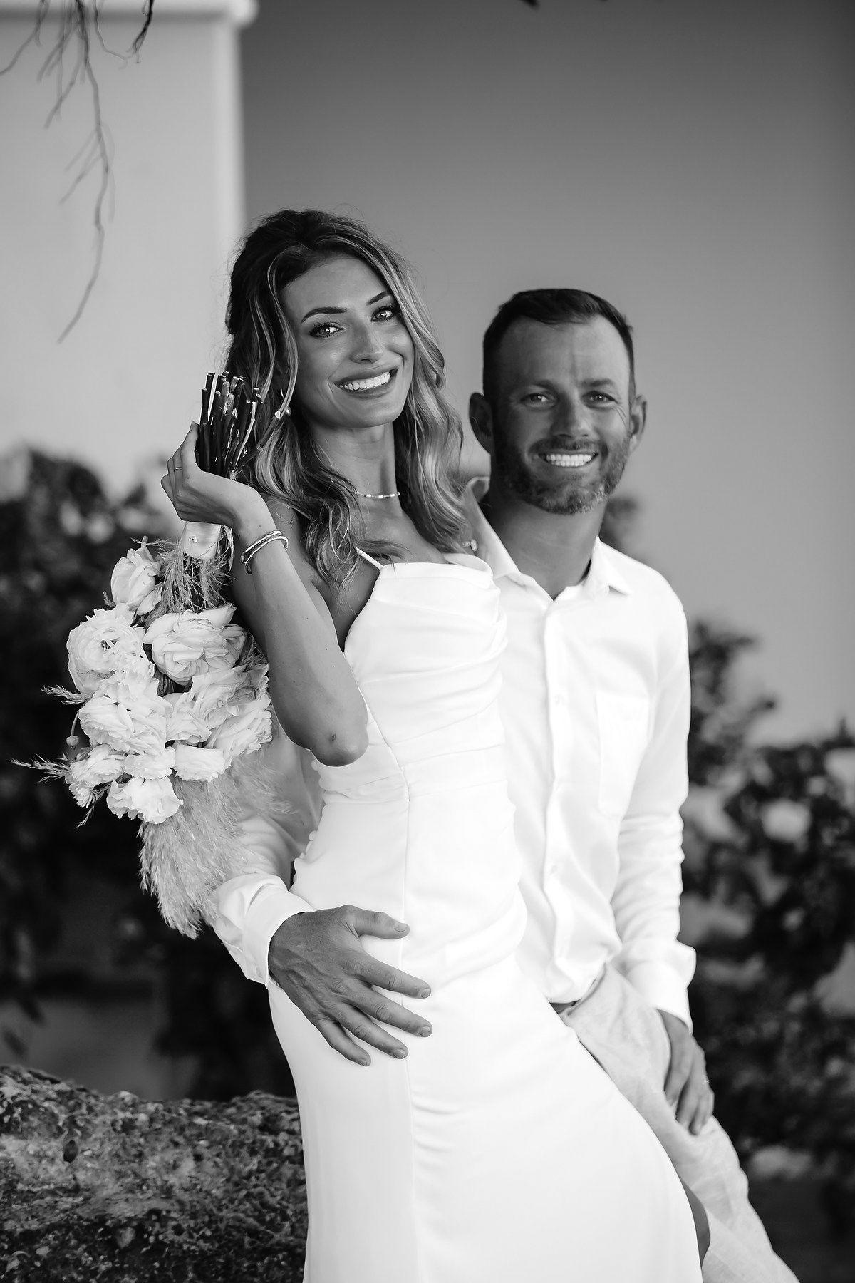 A black and white image of a couple posing together outdoors, with the woman holding a bouquet of flowers.